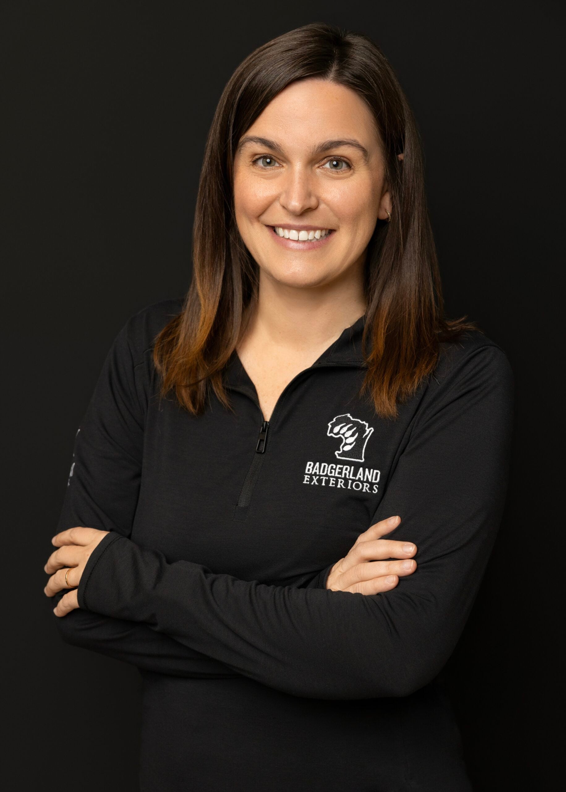 A woman with straight brown hair and a friendly smile stands with arms crossed, wearing a black zip-up shirt with the Baggerland Exteriors logo, against a plain black background.
