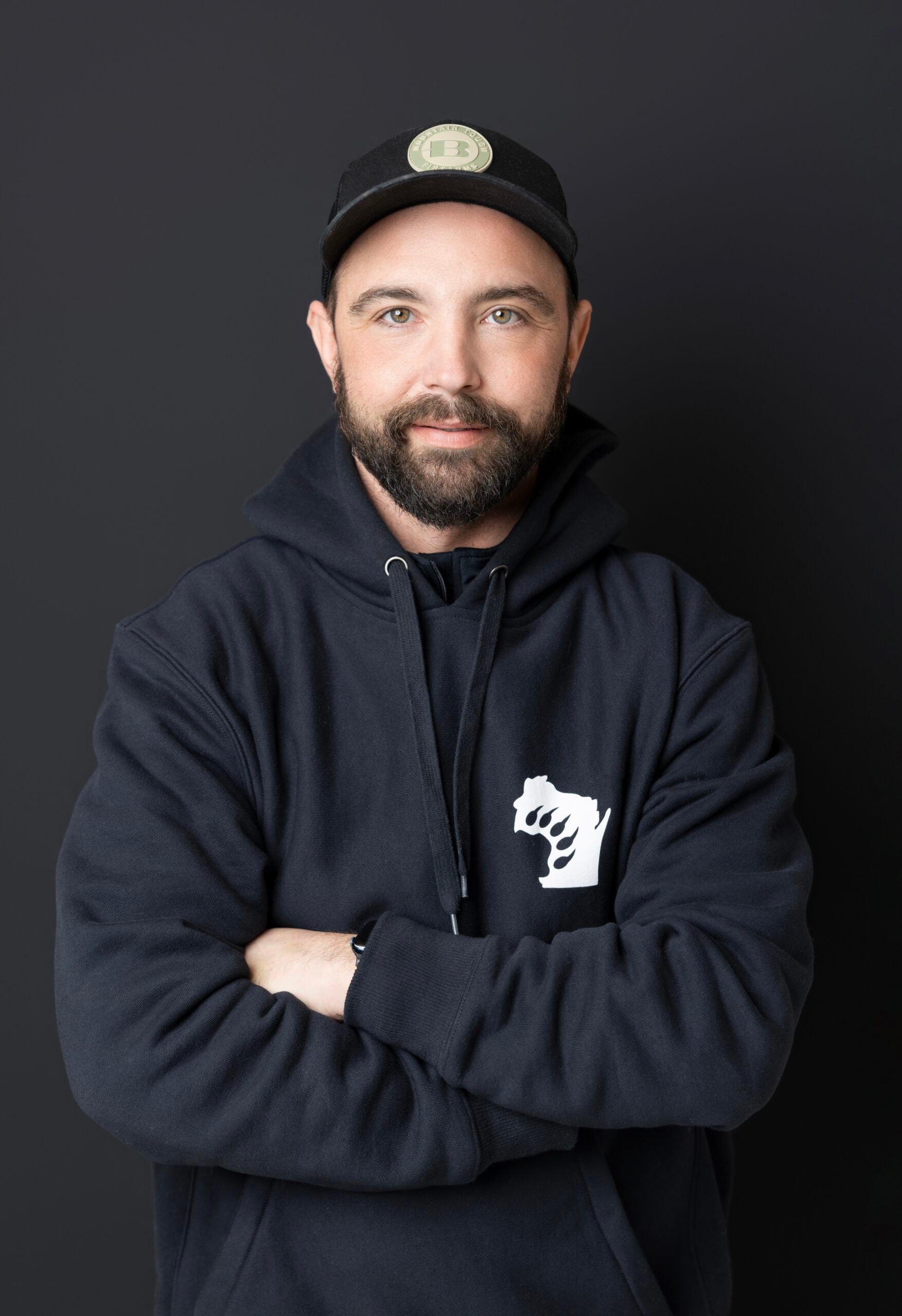 A man with a beard wearing a black cap and black hoodie with a graphic of a raised fist stands against a dark background with his arms crossed, looking at the camera.