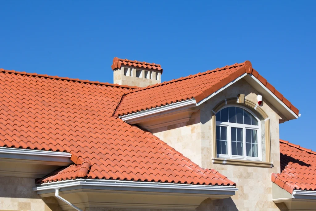 A house with beige stone walls and a red tiled roof showcases one of the timeless types of roofing against a clear blue sky. It features an arched window, a chimney, and white gutters.