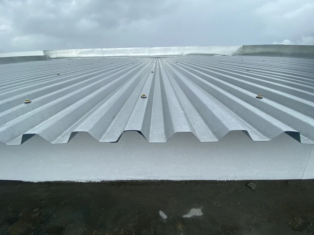 A close-up view of a corrugated metal roof, one popular among various types of roofing, with a slightly overcast sky in the background. The roof has evenly spaced ridges and a few visible fasteners securing the panels.