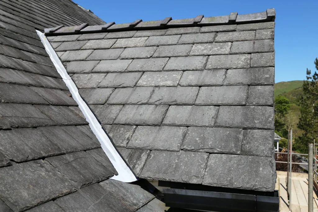 Slate roof with rectangular grey tiles, a prime example of popular types of roofing, features white metal flashing on the seam. The tiles have a slightly weathered appearance set against a clear blue sky and a touch of greenery in the background.
