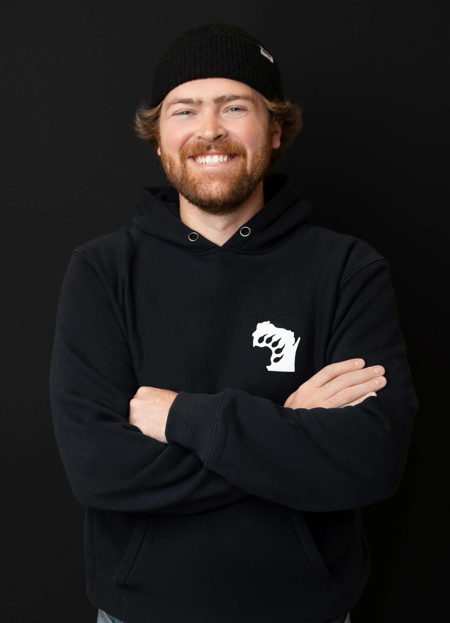A smiling man with a beard and mustache wears a black beanie and a black hoodie with a white fist logo, standing with his arms crossed against a solid black background.