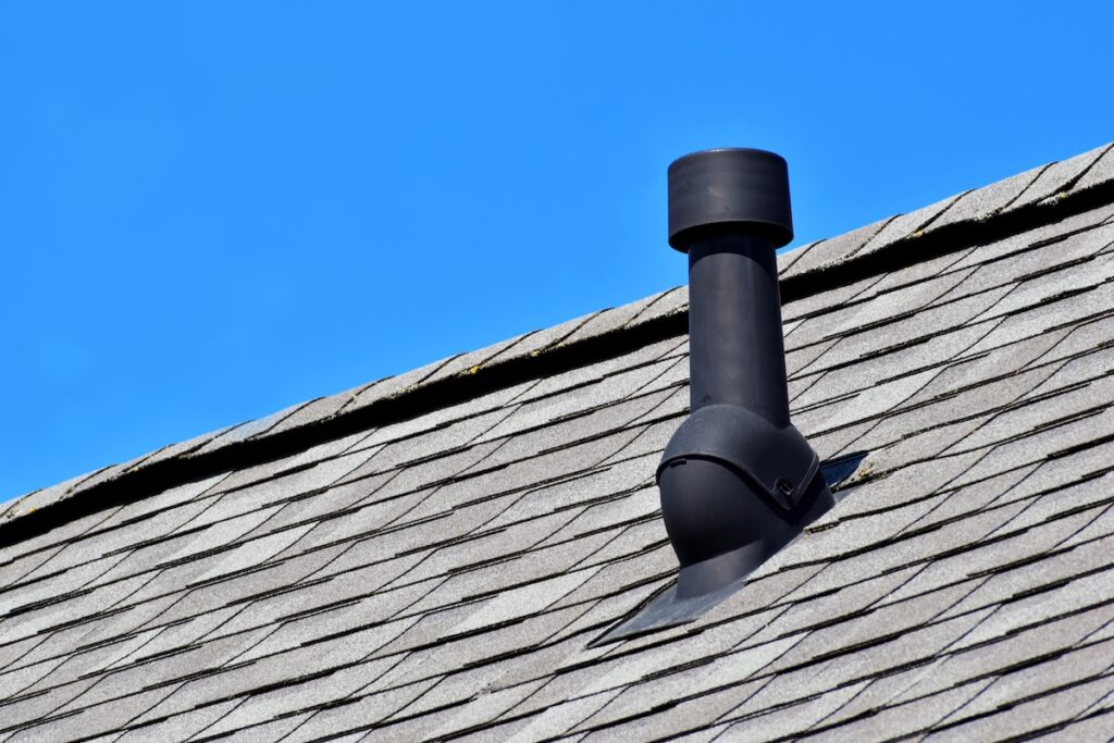 A close-up of a gray shingled roof with a black metal vent pipe extending upward against a clear blue sky, showcasing proper roof ventilation.