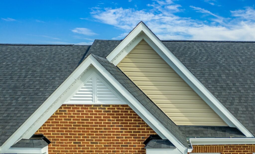 Close-up view of a house roof with two intersecting gables and a distinct roof pitch, featuring dark gray shingles, brick walls, and beige and white siding, set against a blue sky with scattered clouds.
