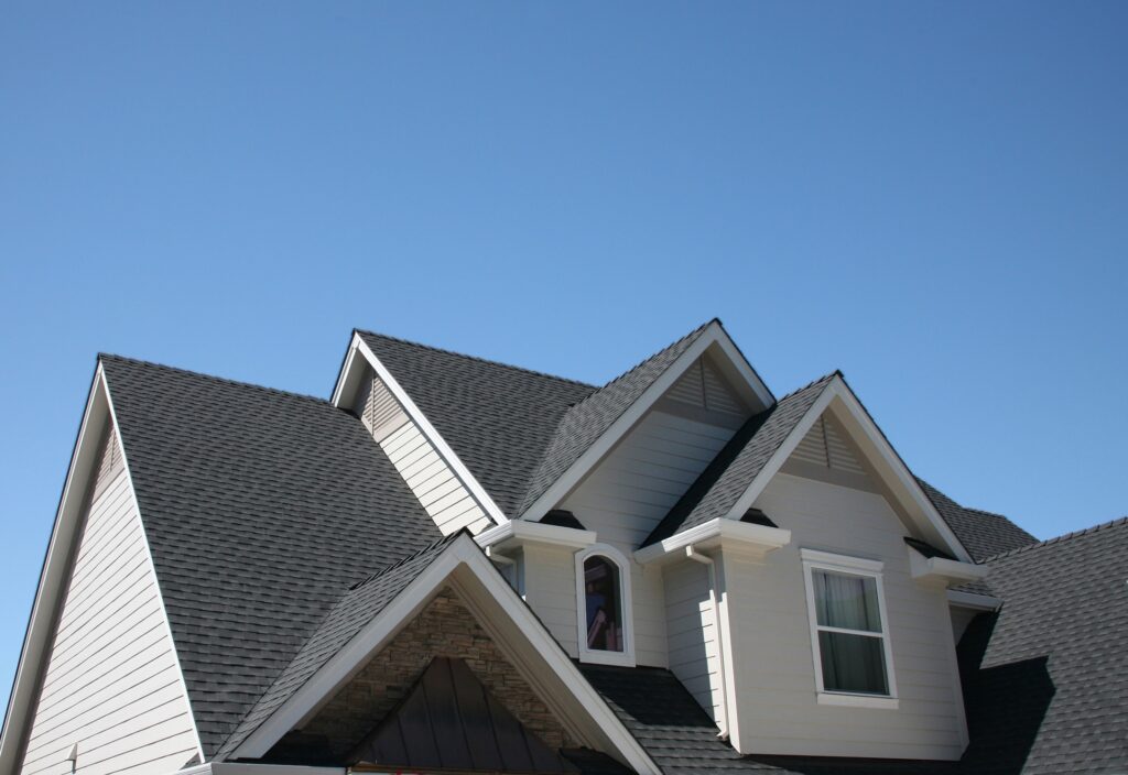 Close-up of a modern house roof with multiple gables and dark gray shingles against a clear blue sky. The house exterior is light-colored with white trim and features windows on the upper floor.