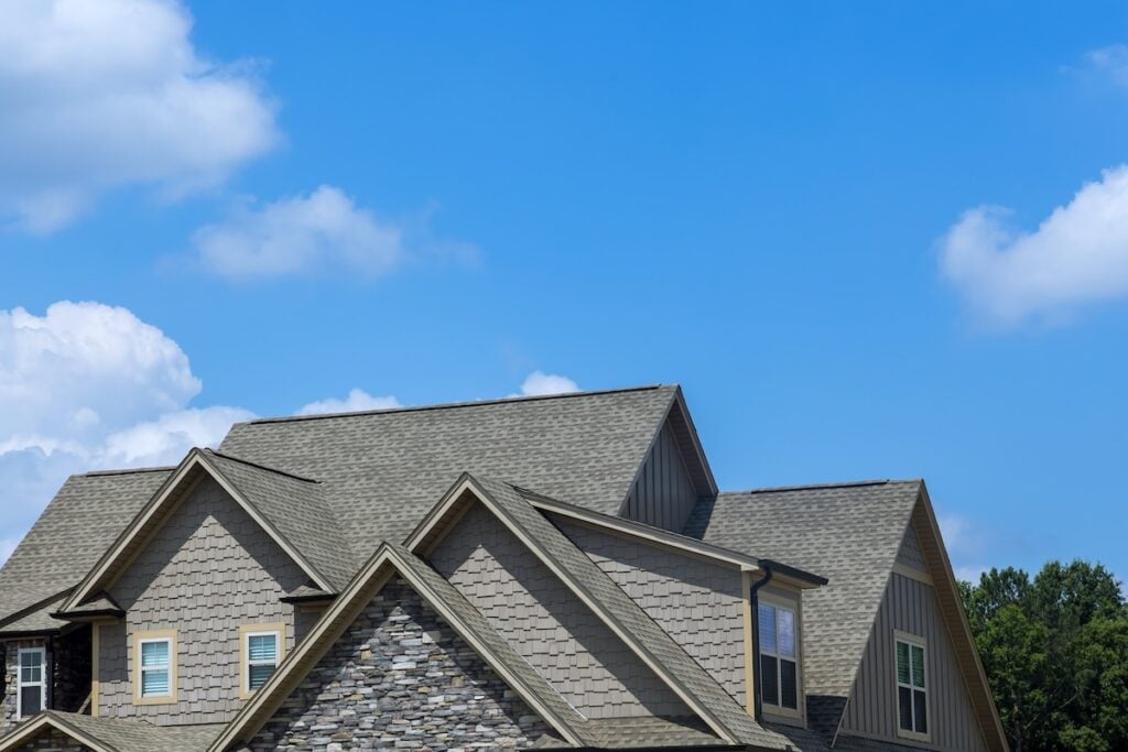 The rooftops of a modern suburban house with gray shingles and exterior stonework are seen against a bright blue sky with scattered clouds. Trees are visible in the background.