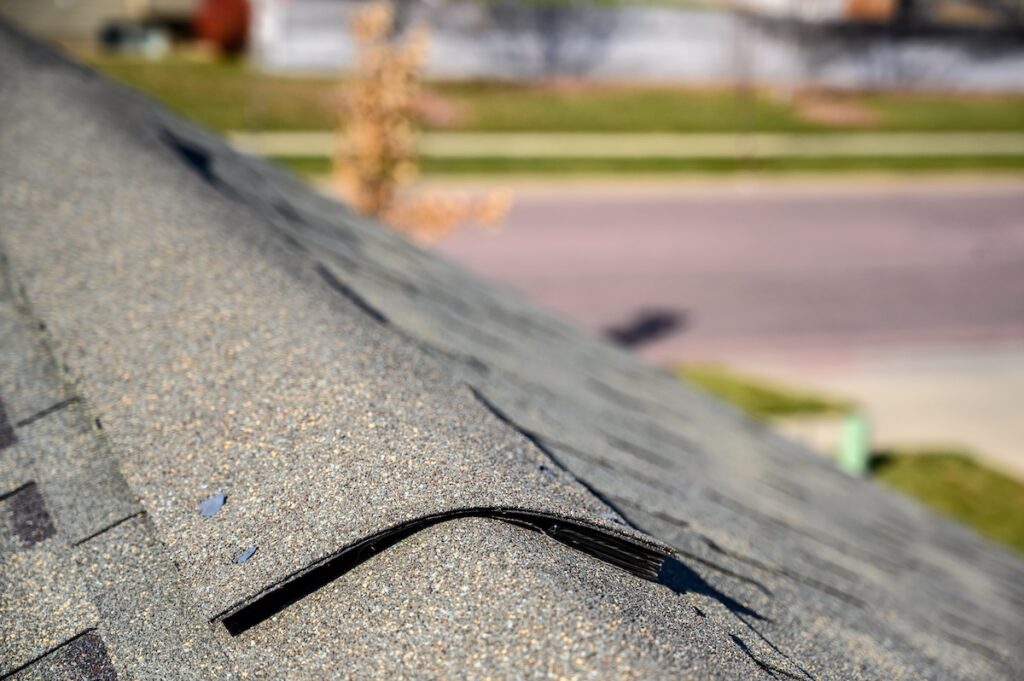 Close-up of an asphalt shingle roof with a loose or damaged ridge cap shingle sticking up, indicating potential roof damage. A blurred street and lawn are visible in the background.