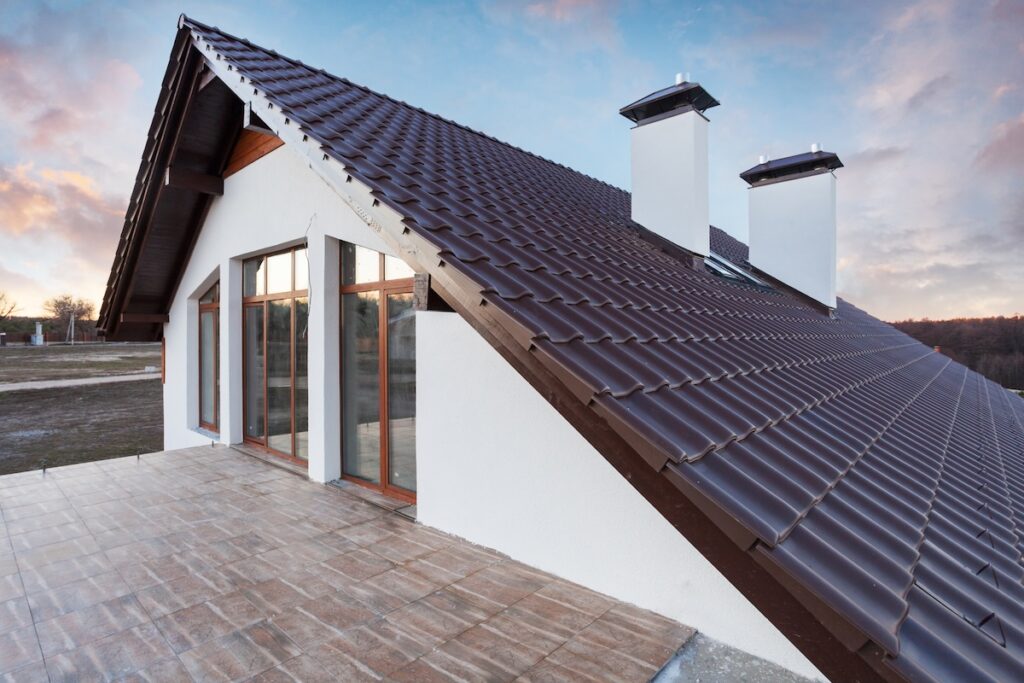 A modern house with a steep roof pitch featuring brown tiles, white walls, large windows, and a spacious tiled terrace, set against a scenic outdoor background at sunset.