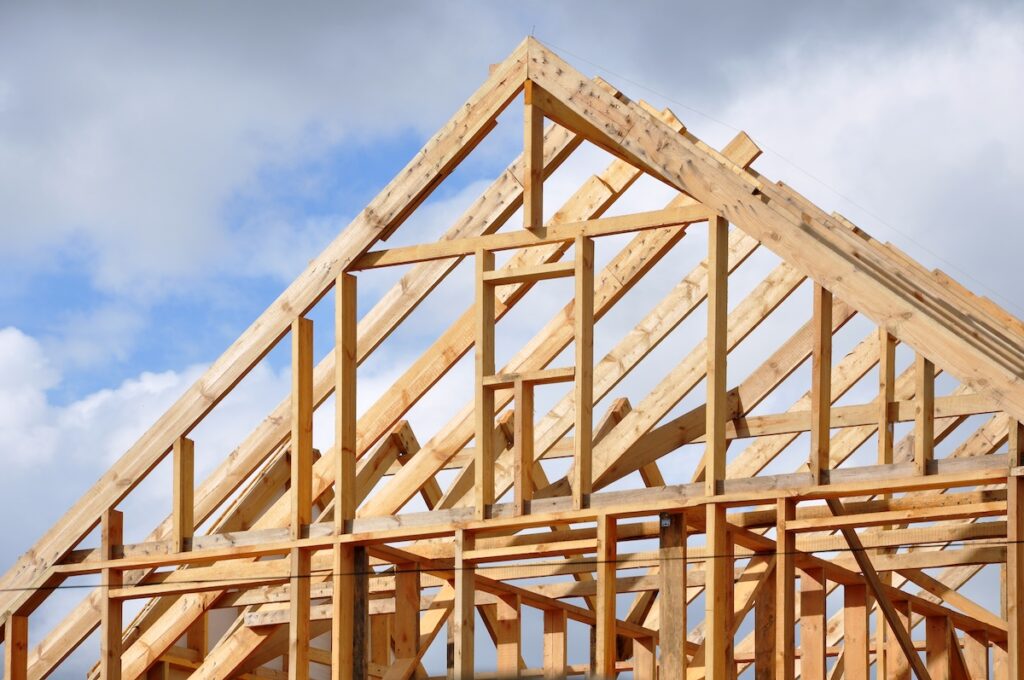 The image shows the wooden frame of a house under construction, with visible beams and roof trusses set against a partly cloudy sky in the background.