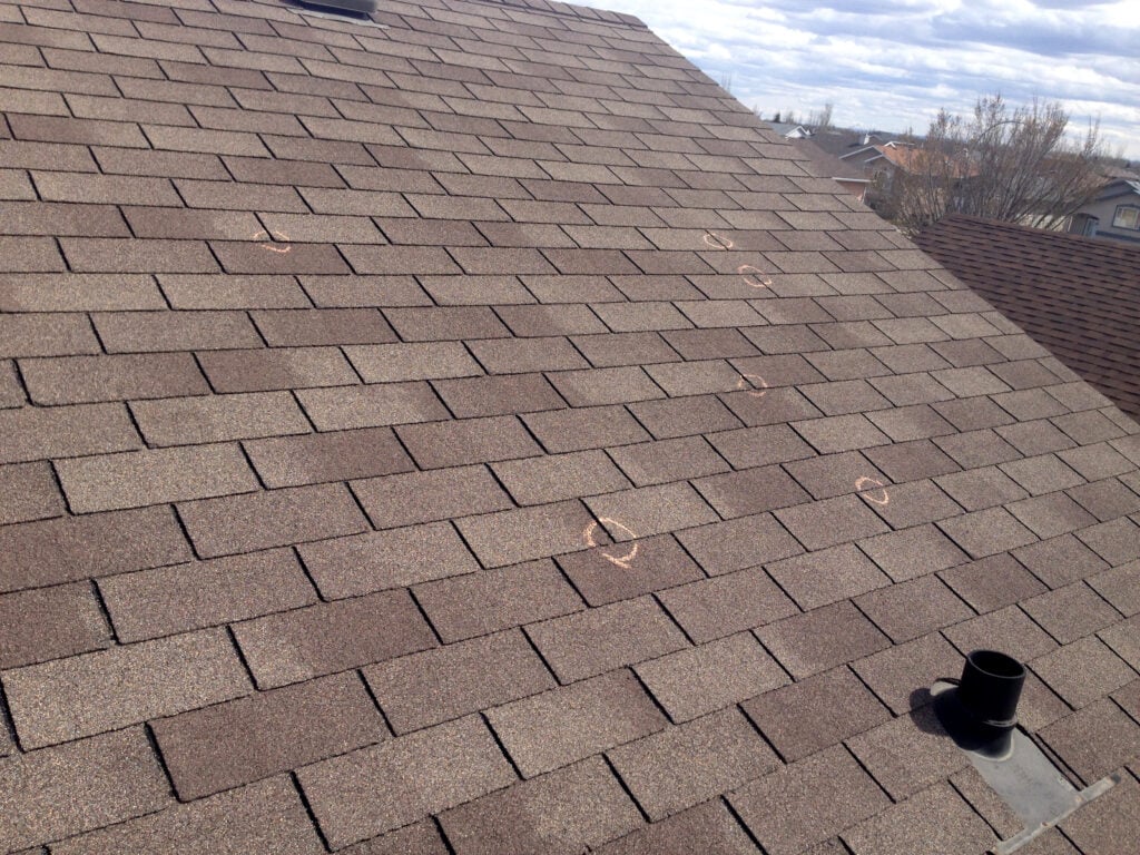 A brown shingled roof displays several chalk circles marking areas from a recent hail damage roof inspection under a cloudy sky, with neighboring rooftops and trees visible in the background.
