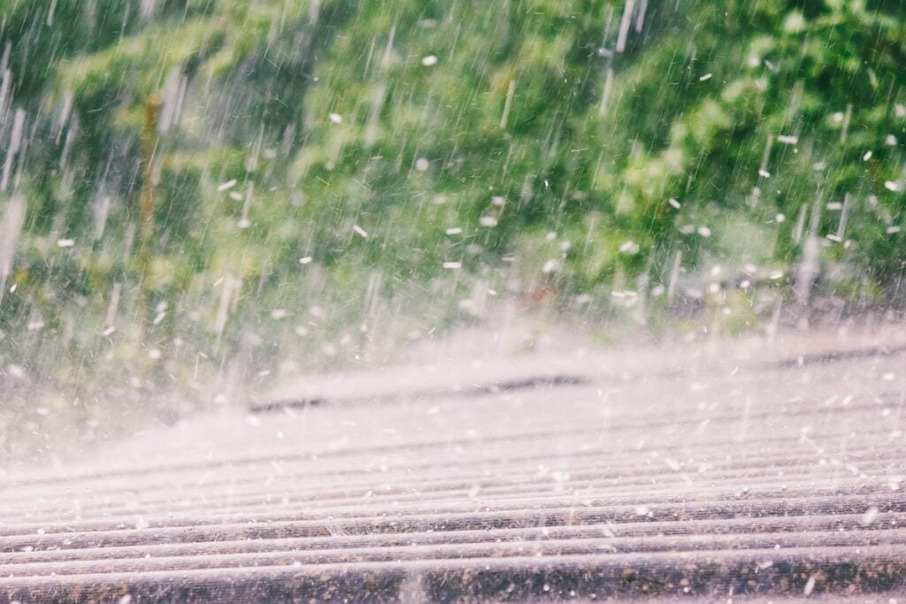 Heavy rain pours down over a rooftop, with raindrops visibly splashing. The blurred green foliage in the background enhances the sense of motion and wet weather—ideal conditions for a hail damage roof inspection.