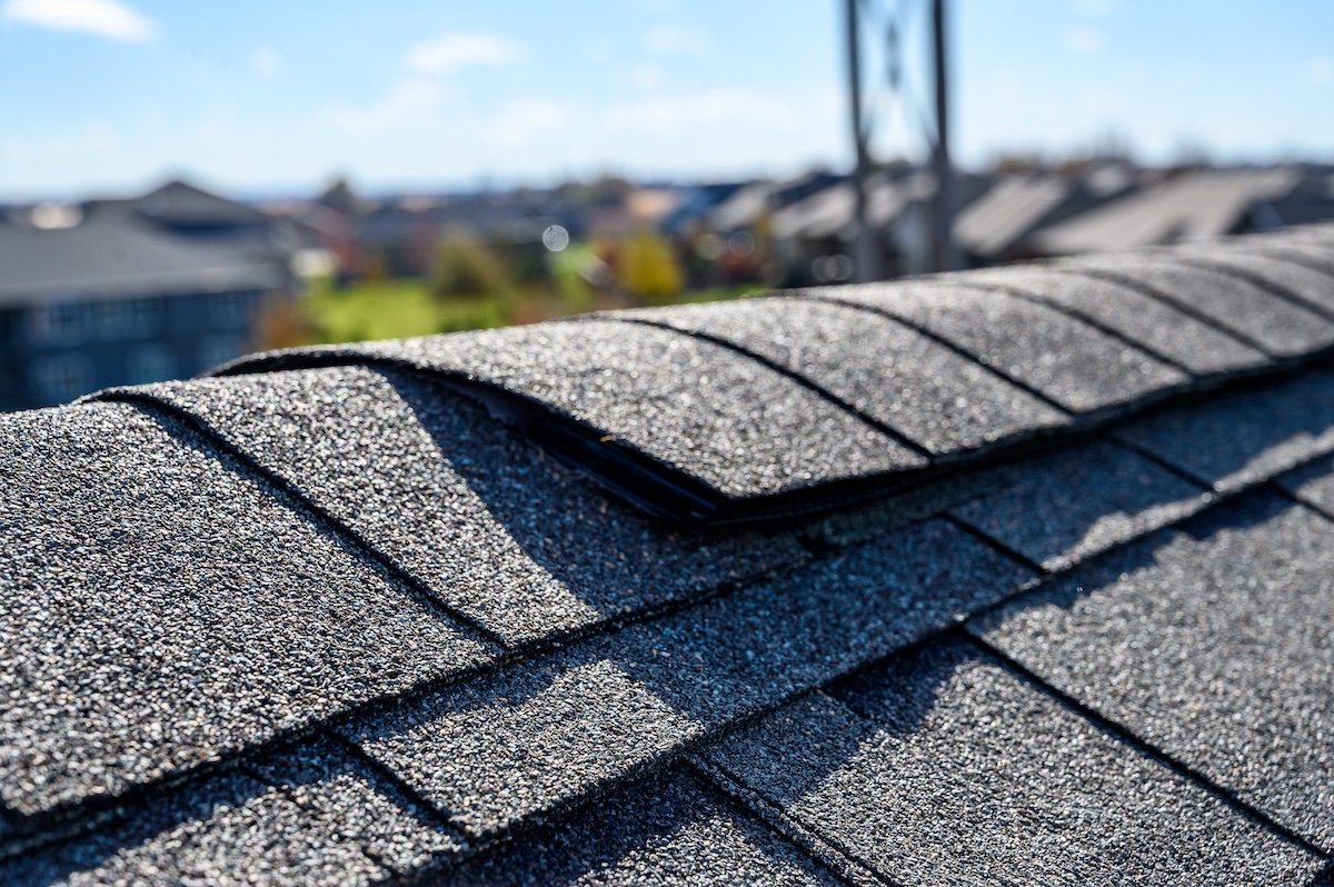 Close-up of an asphalt shingle roof with some shingles lifted or damaged, including visible ridge cap shingles, with a blurred background of other buildings and a blue sky.
