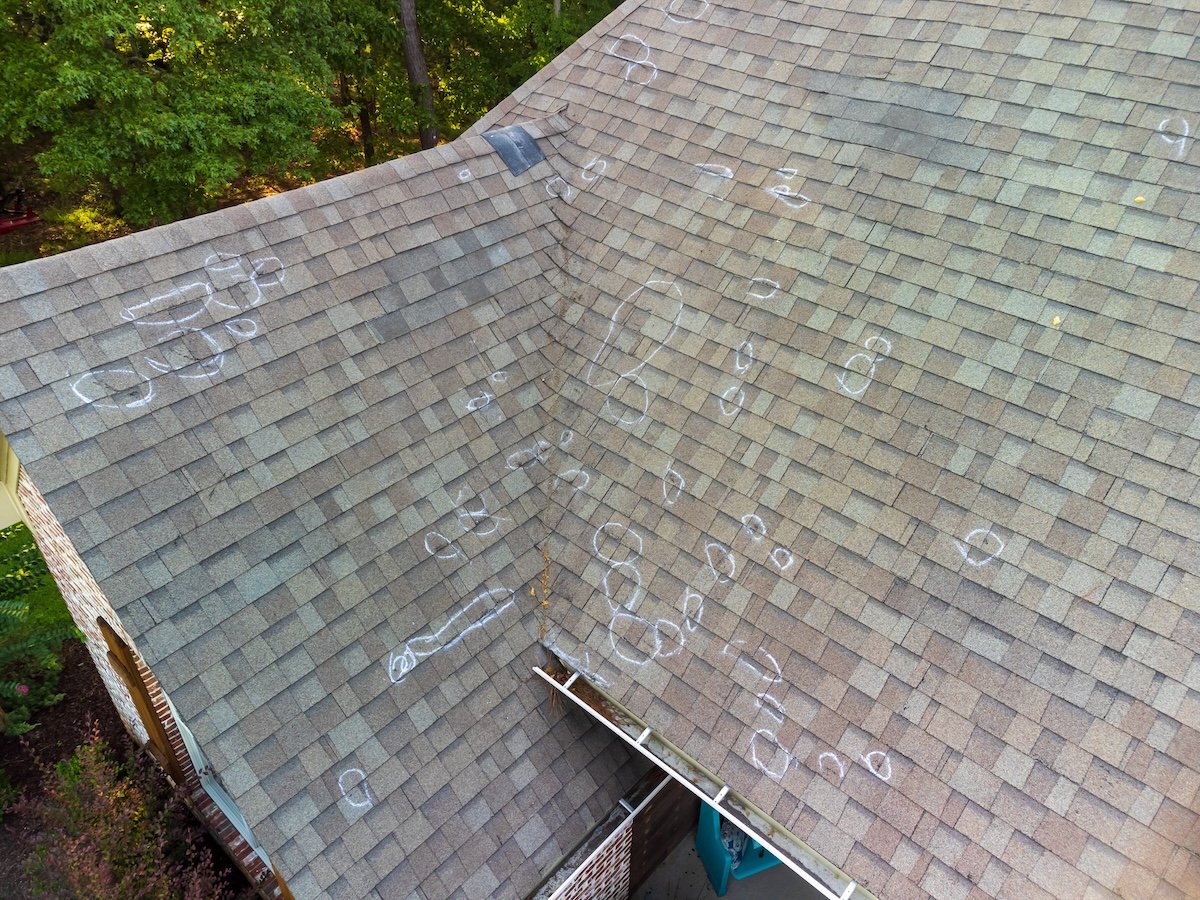 A house roof with many chalk circles marking hail damage on the shingles, surrounded by trees—evidence of a recent hail damage roof inspection.