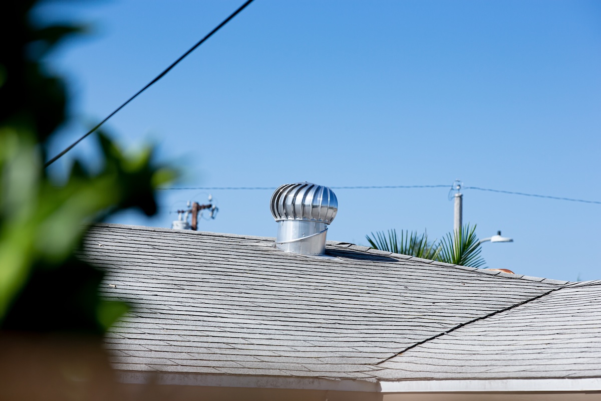 A metal roof vent designed for roof ventilation sits on top of a shingled roof under a clear blue sky, with power lines and a few trees in the background.
