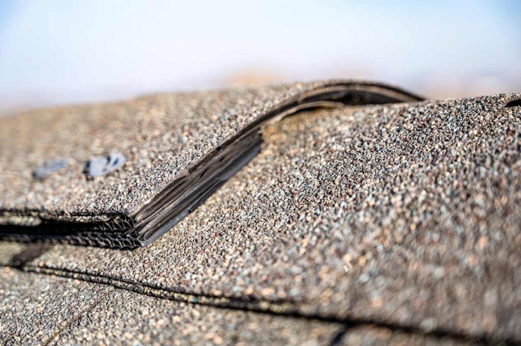 Close-up view of overlapping asphalt roof shingles, with one shingle slightly lifted to reveal the rough, textured surface and granular details—highlighting the quality and installation of ridge cap shingles. The image is focused on the roofing material.