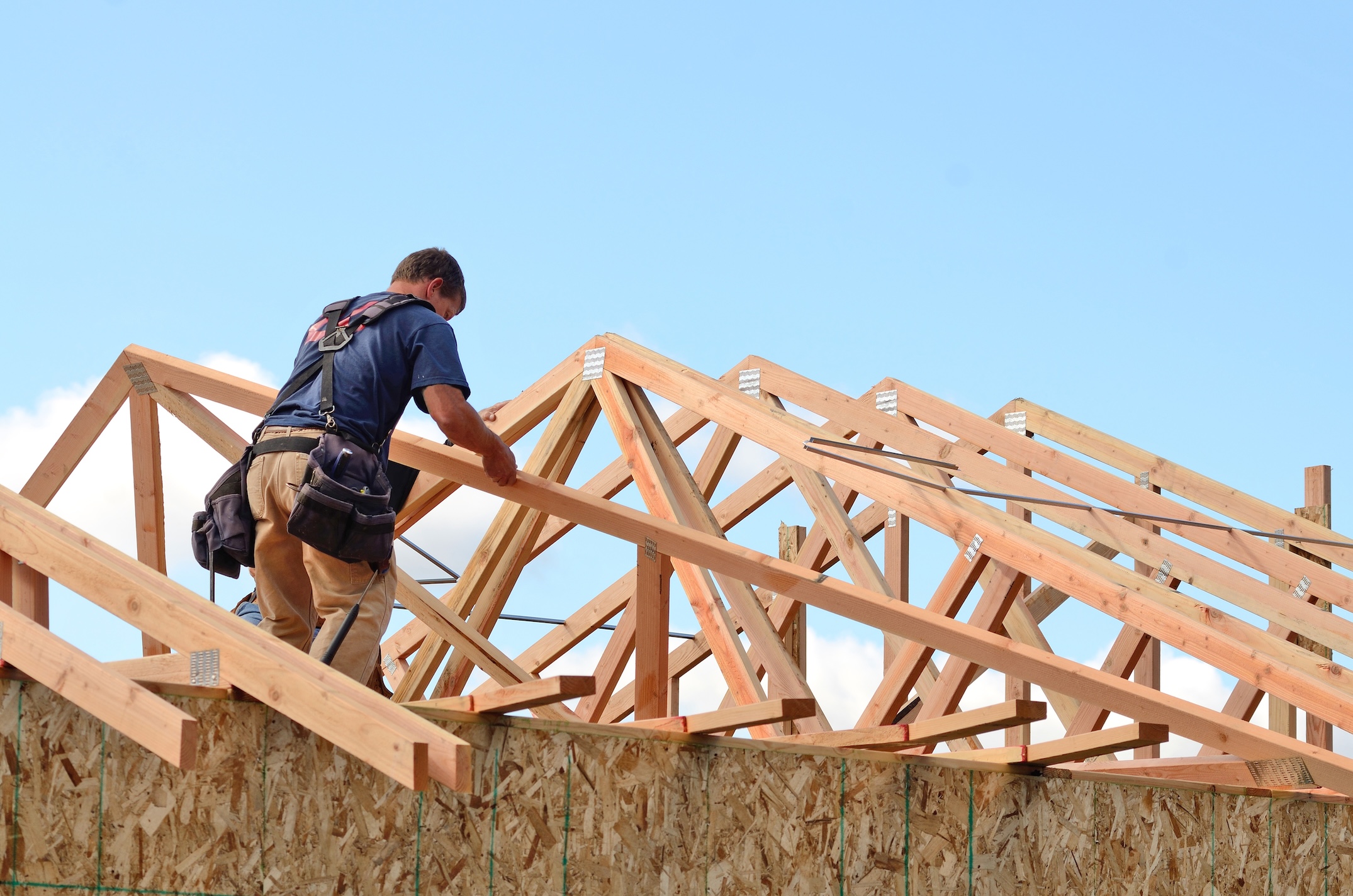 A construction worker wearing a tool belt works on assembling roof trusses of a building under construction against a clear blue sky.