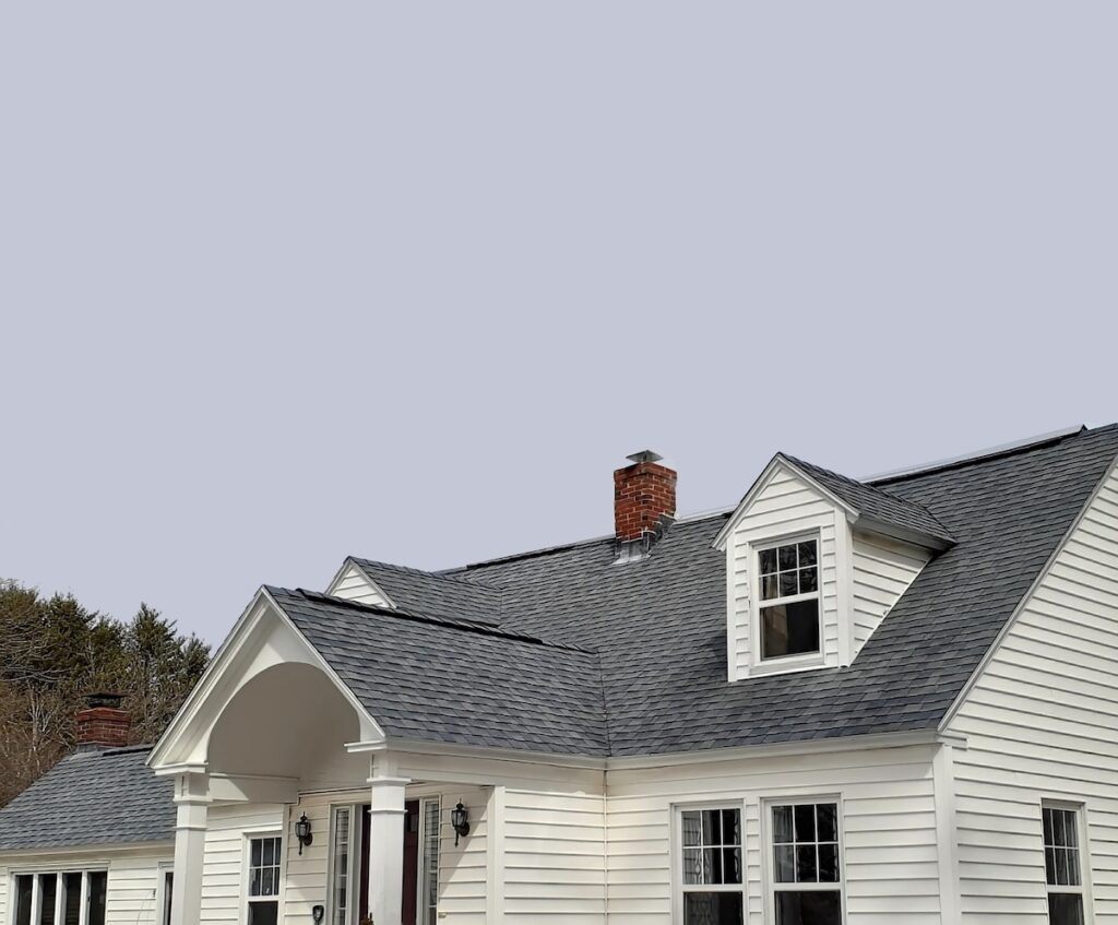 A white house with gray shingles, a prominent front porch, dormer windows, and a red brick chimney, set against a clear, pale sky. Some trees are partially visible to the left.