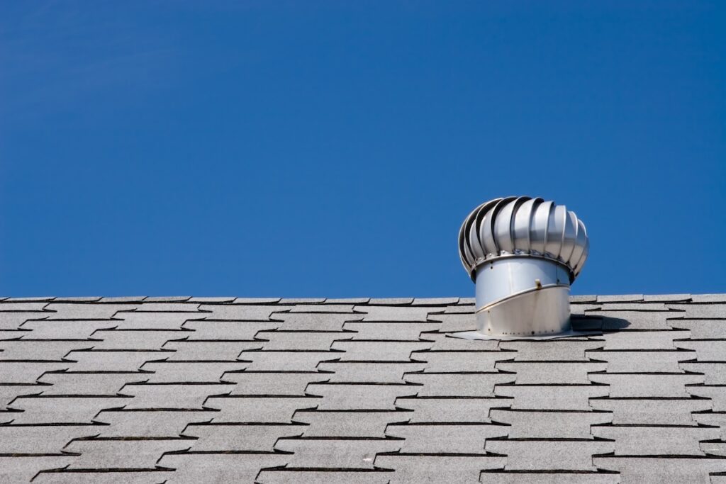 A silver metal roof vent, designed for roof ventilation, sits on a shingled rooftop under a clear blue sky. The turbine-style vent is positioned near the right side of the image.
