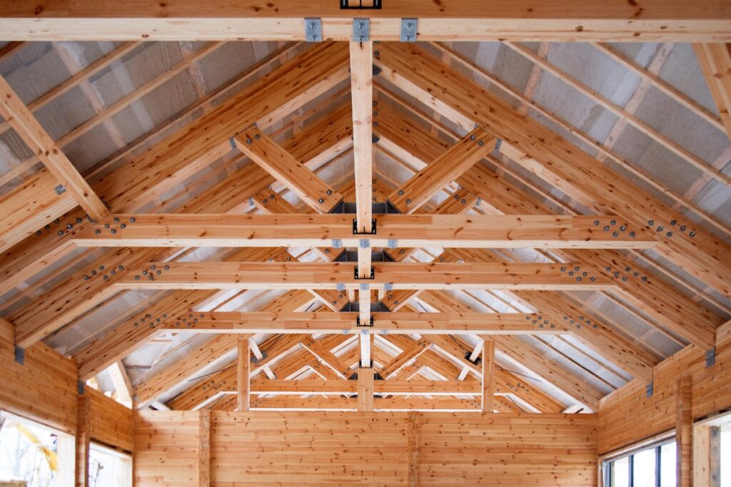 Interior view of a wooden building under construction, showing exposed timber beams, roof trusses, and metal connectors forming the ceiling structure.