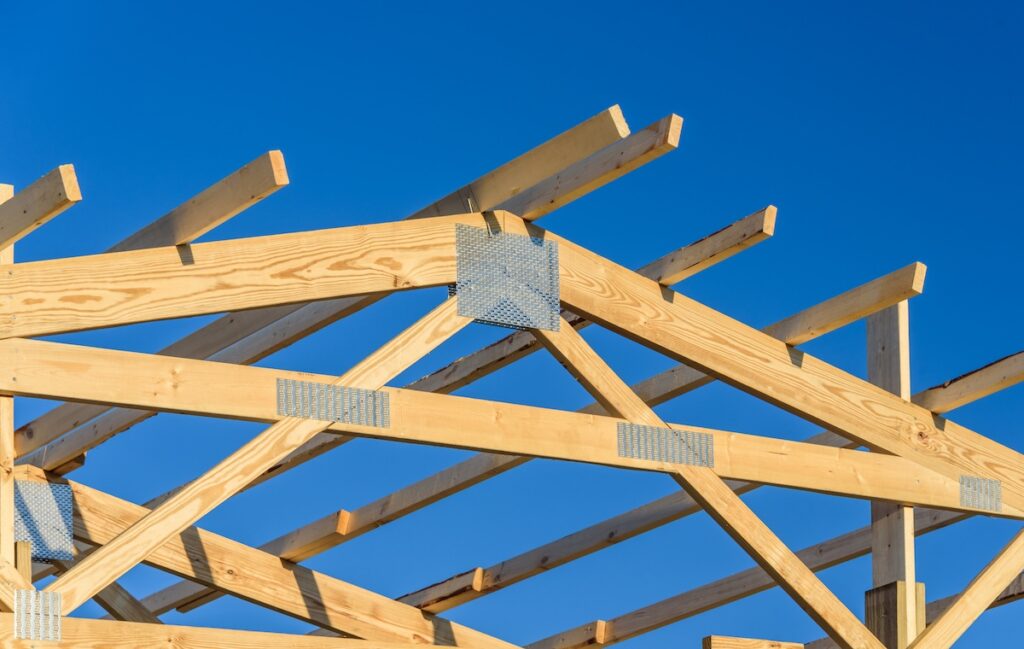 Partially constructed roof trusses form the wooden structure of a building, with light-colored beams and metal connectors, set against a clear blue sky.