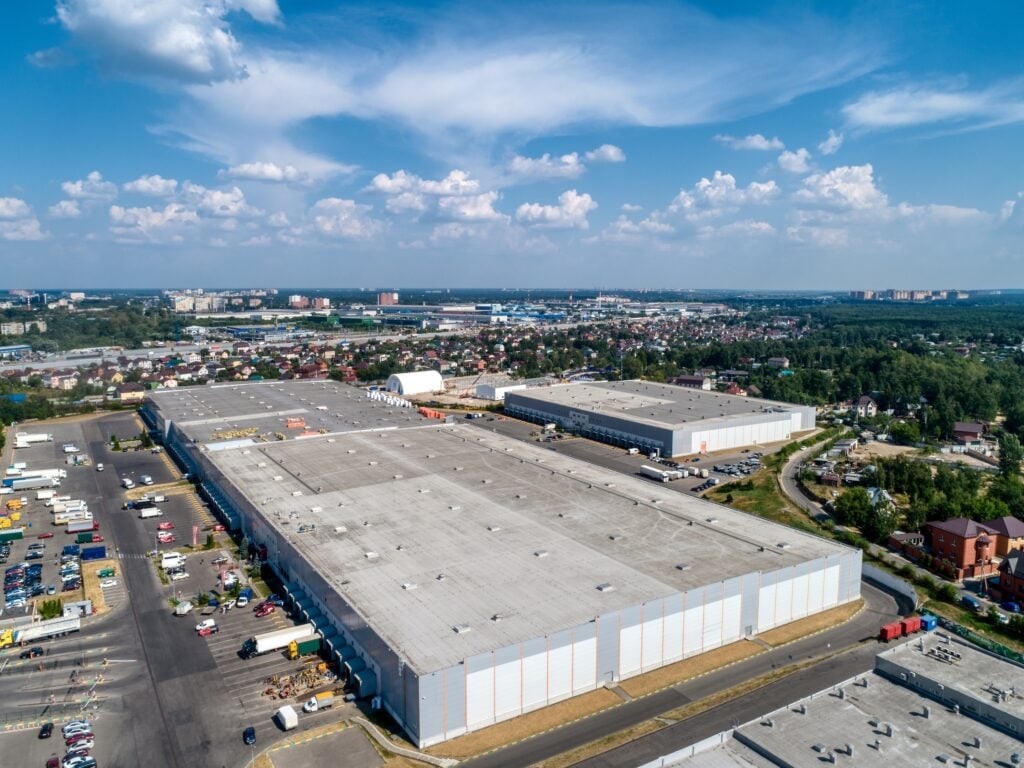 Aerial view of a large industrial warehouse complex showcasing various commercial roof types, surrounded by parking lots, trucks, and smaller buildings, with a cityscape and greenery in the background under a partly cloudy sky.