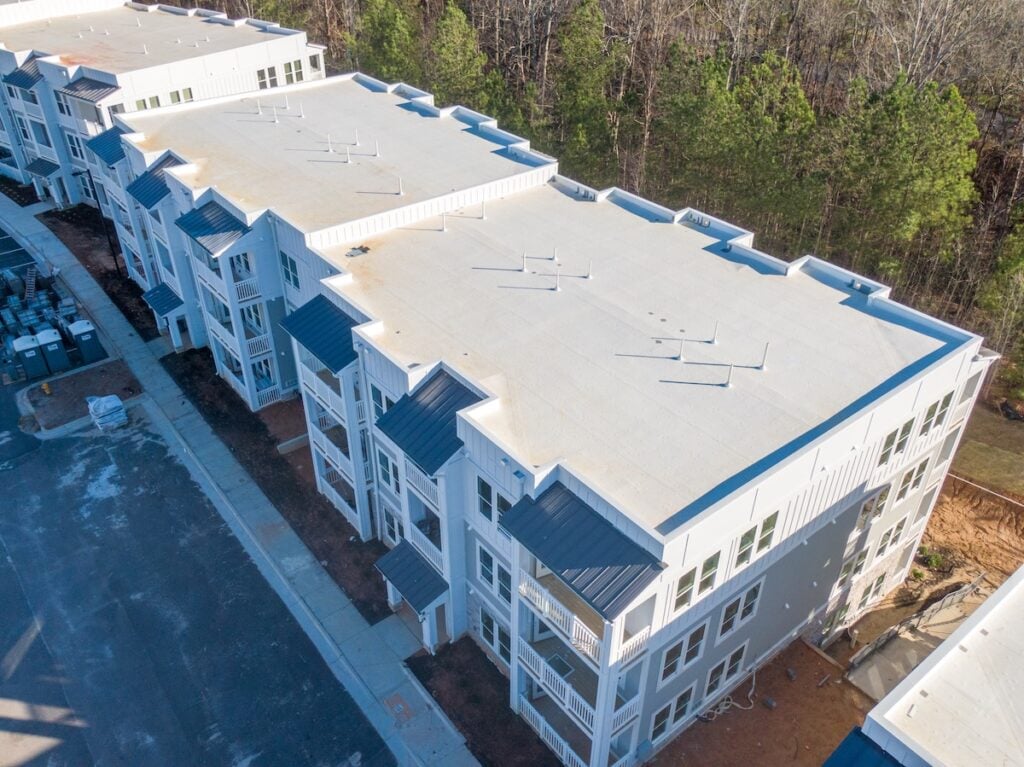Aerial view of a modern, three-story apartment building with a white commercial roof type and exterior, surrounded by trees and an unfinished area scattered with dirt and construction materials. commercial roof types
