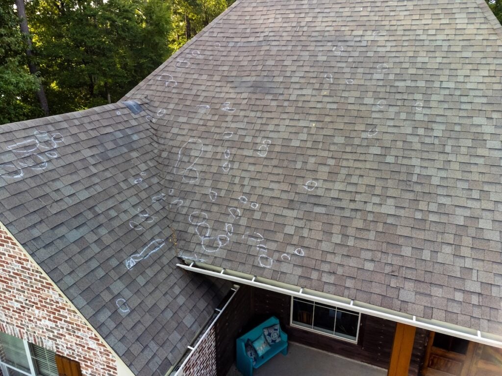 Aerial view of a house roof with numerous chalk circles marking spots on the shingles, possibly indicating types of roof damage or inspection points. Trees are visible in the background.