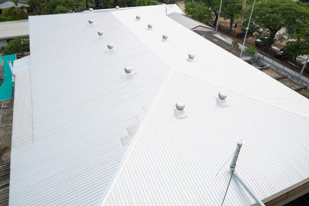A large white metal commercial roof, one of the popular commercial roof types, is shown from above with multiple ventilators. Trees, buildings, and a street border the roof’s perimeter.