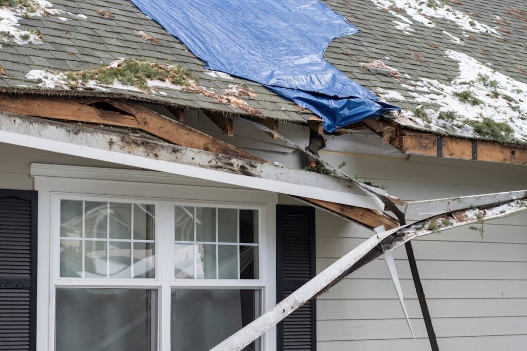A house roof displays types of roof damage, with missing shingles, exposed wood, and a blue tarp. Snow covers part of the surface, while a bent gutter hangs near a window with black shutters.