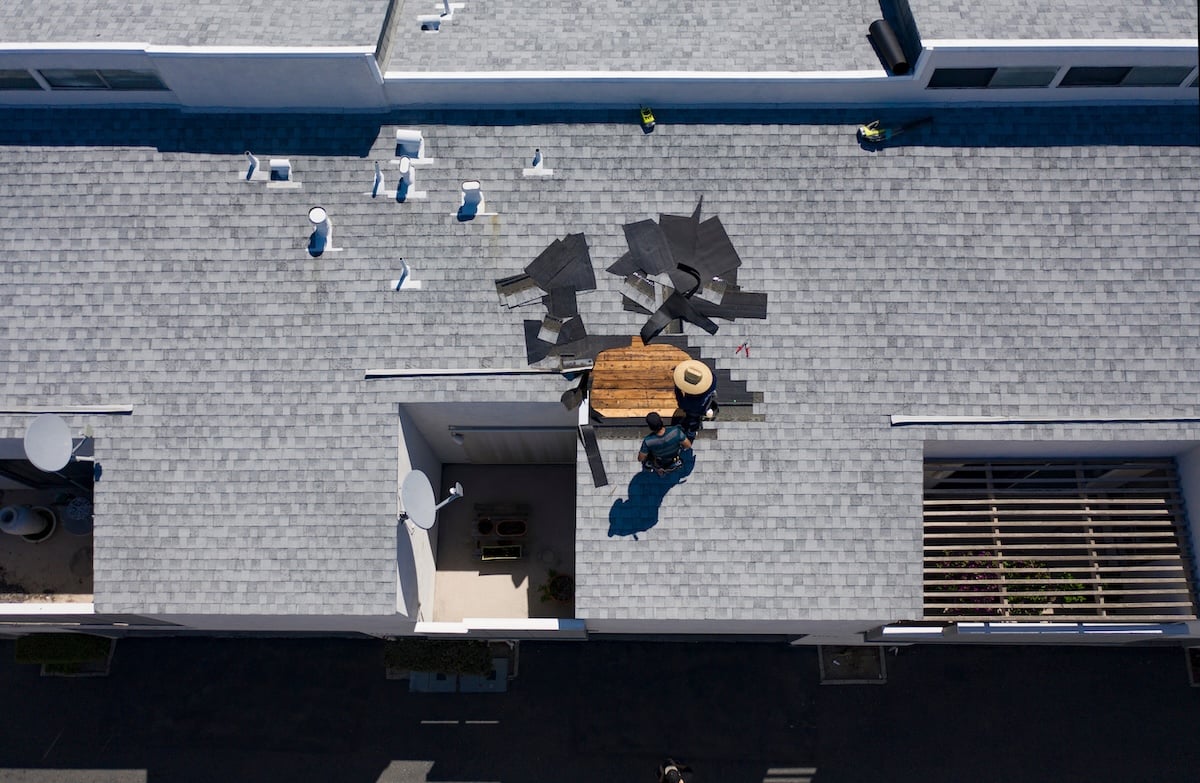 Aerial view of two workers repairing a gray shingle roof, addressing various types of roof damage. Roofing materials are spread out around them. Shadows indicate a sunny day, highlighting vents and skylights on the building.