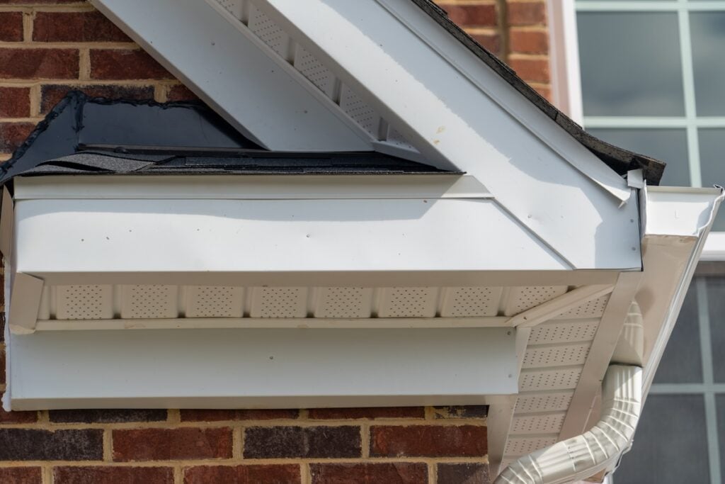 Close-up of a house’s roof corner showing white gutters, soffit, fascia, drip edge, and downspout against a brick wall. Part of a window is visible on the right side of the image. drip edge