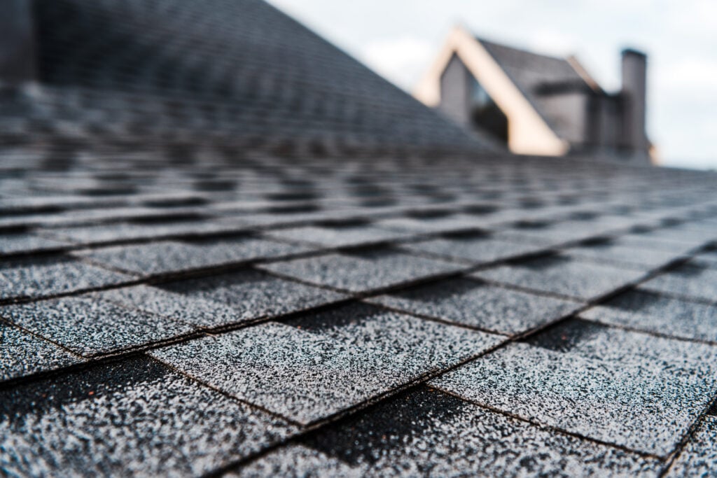 Close-up view of gray asphalt shingles showing slight granular loss on roof, with a house and chimney blurred in the background under a partly cloudy sky.