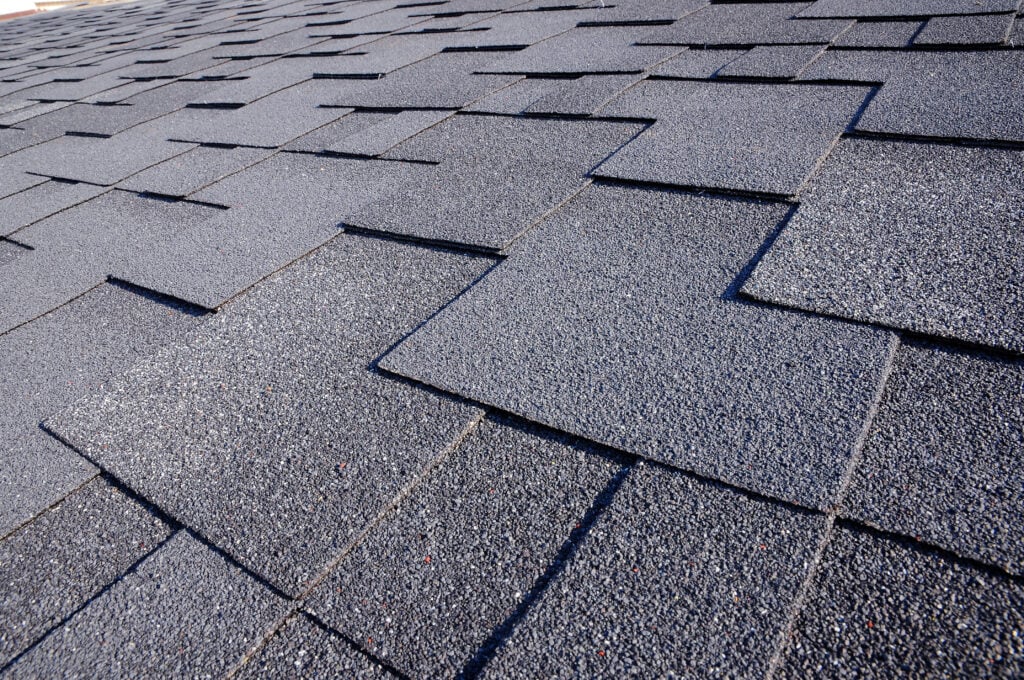 Close-up view of overlapping gray asphalt roof shingles arranged in a uniform pattern, highlighting their texture and areas of granular loss on the roof.