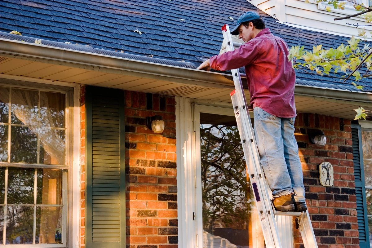 A person stands on a ladder performing gutter cleaning on a brick house. Wearing jeans, a red long-sleeve shirt, and a blue cap, they clear leaves from the gutter near the roof and beside the window by the door.