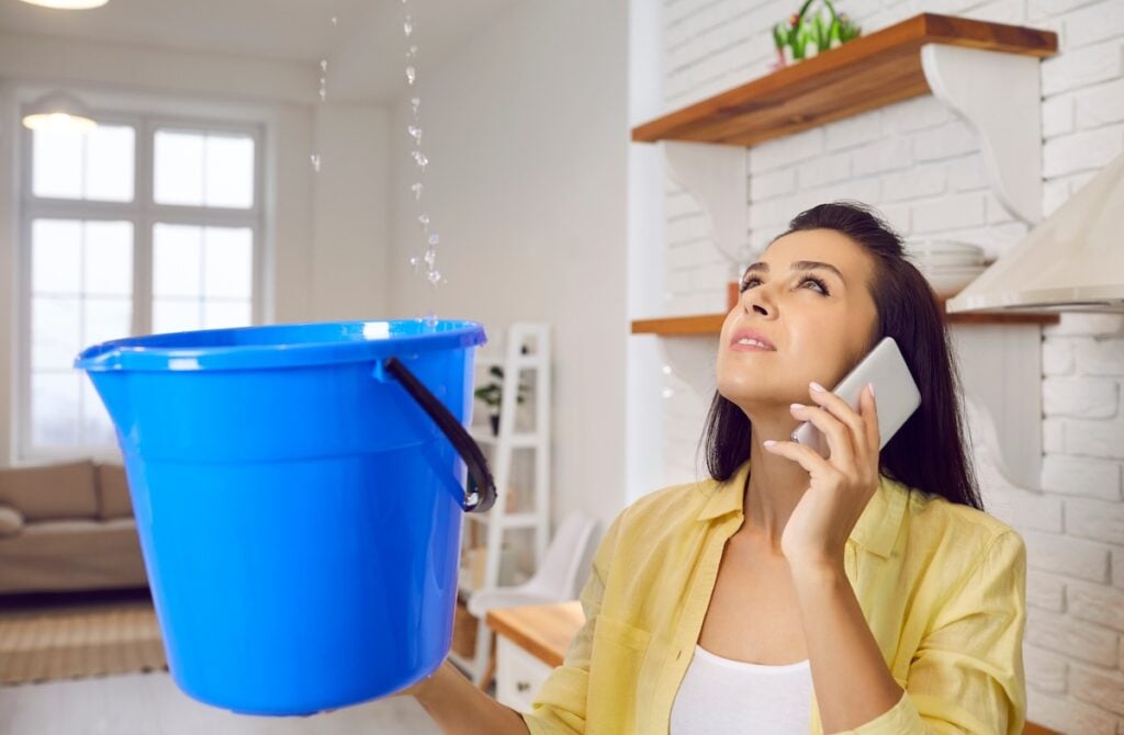 A woman holds a blue bucket under a ceiling leak while talking on her phone, looking up at the dripping water in a home kitchen or living area.
