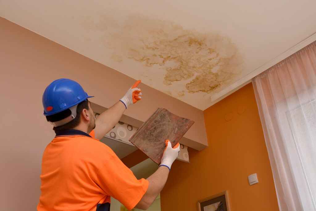 A worker in an orange shirt and blue hard hat points to a large water stain on a ceiling while holding a trowel, indicating damage that needs repair.
