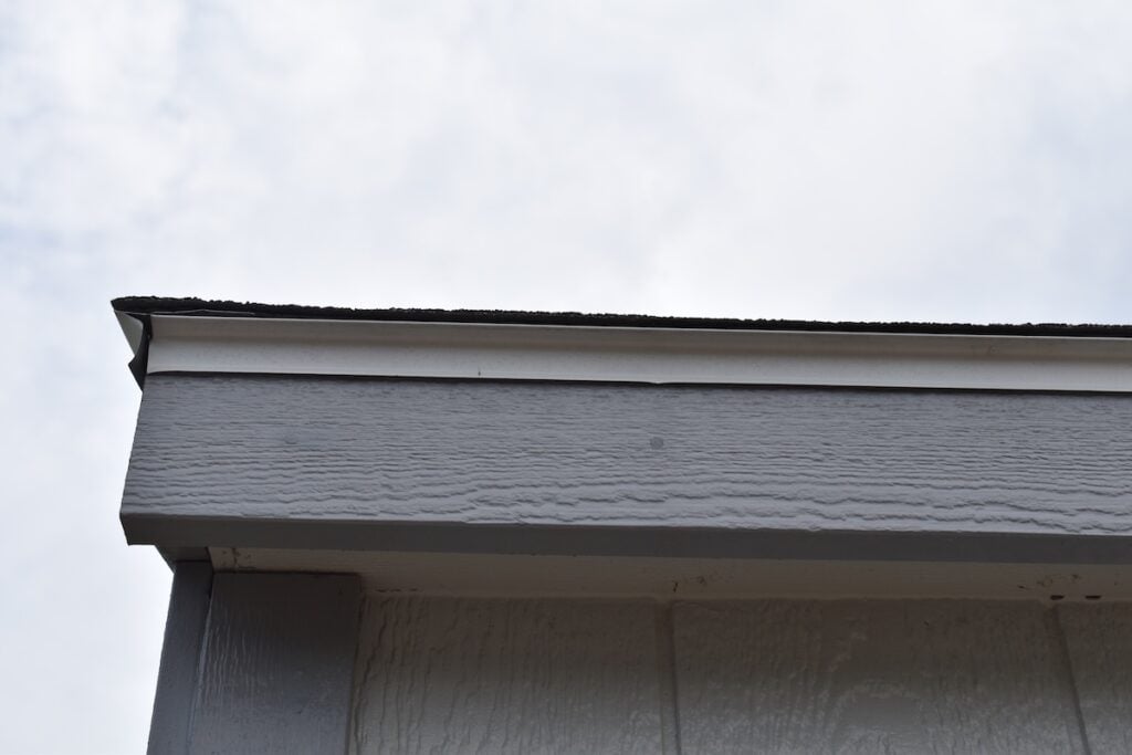 Close-up view of the roof edge and fascia of a gray building against a cloudy sky, showing the siding, trim, drip edge, and roof shingles.