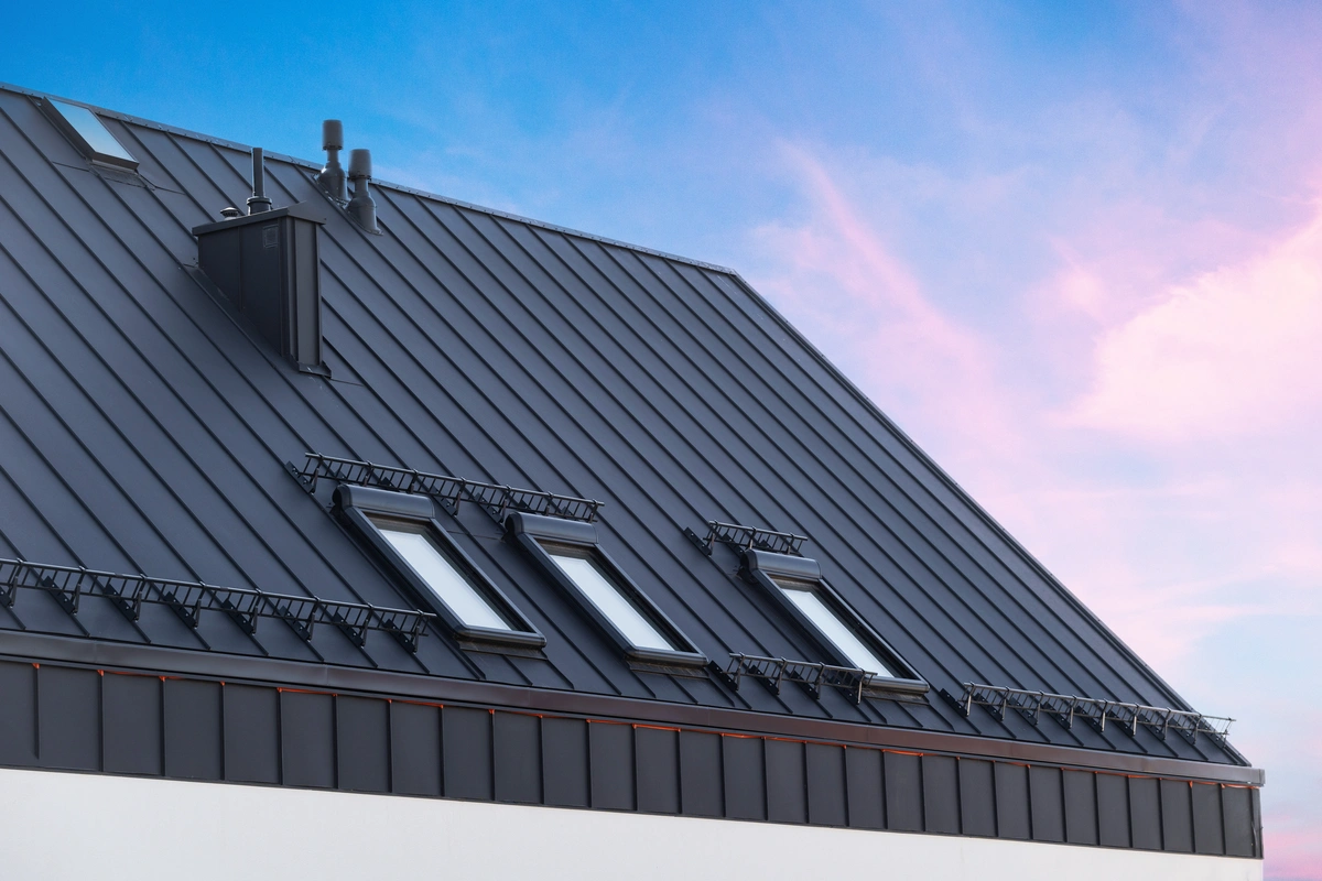 A modern sloped metal roof with four skylight windows and snow guards, set against a blue sky with pink clouds.