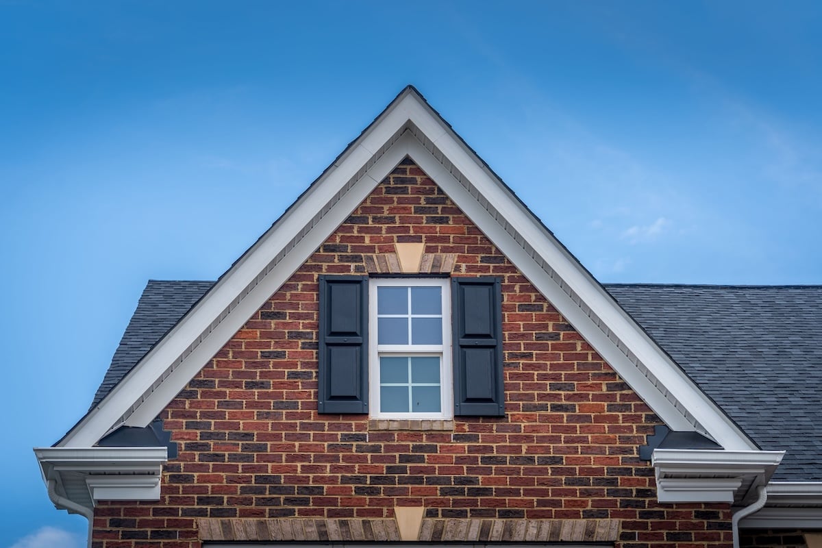 A gable of a brick house with a peaked roof, a white-framed window, and closed black shutters against a clear blue sky.