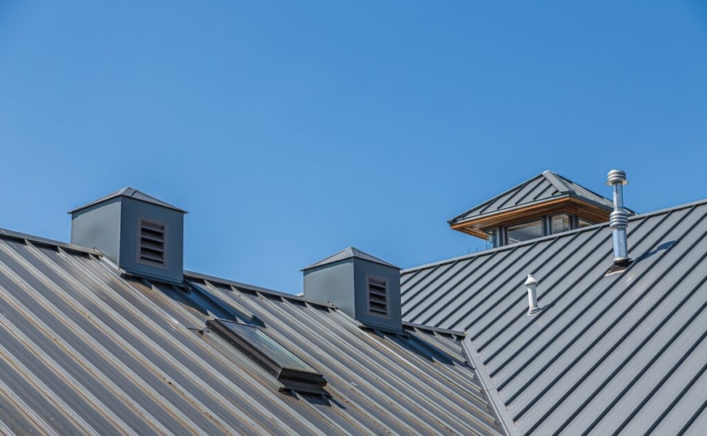 A close-up view of a metal roof with ridges, vents, and a small chimney under a clear blue sky. The roof has two square ventilation structures and a dormer window.