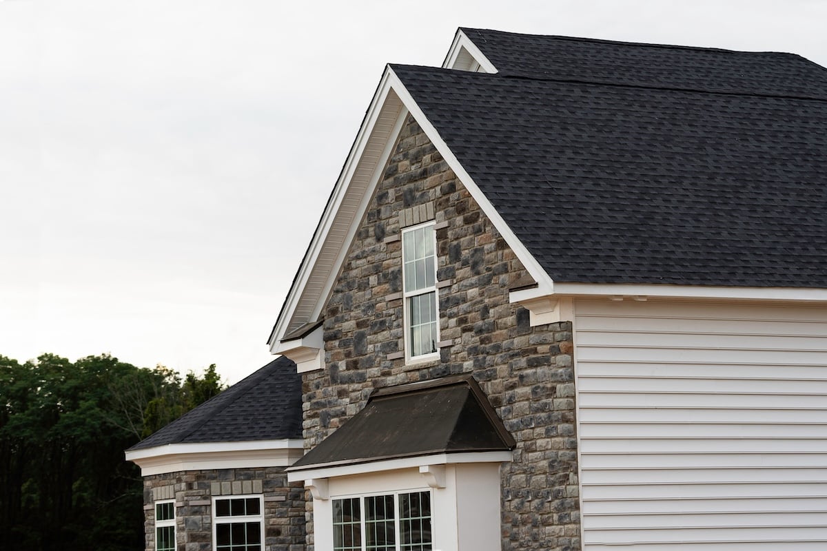A close-up of a modern house exterior featuring gray stone and white siding, peaked roofs with dark shingles, and large windows. Trees are visible in the background under a cloudy sky.