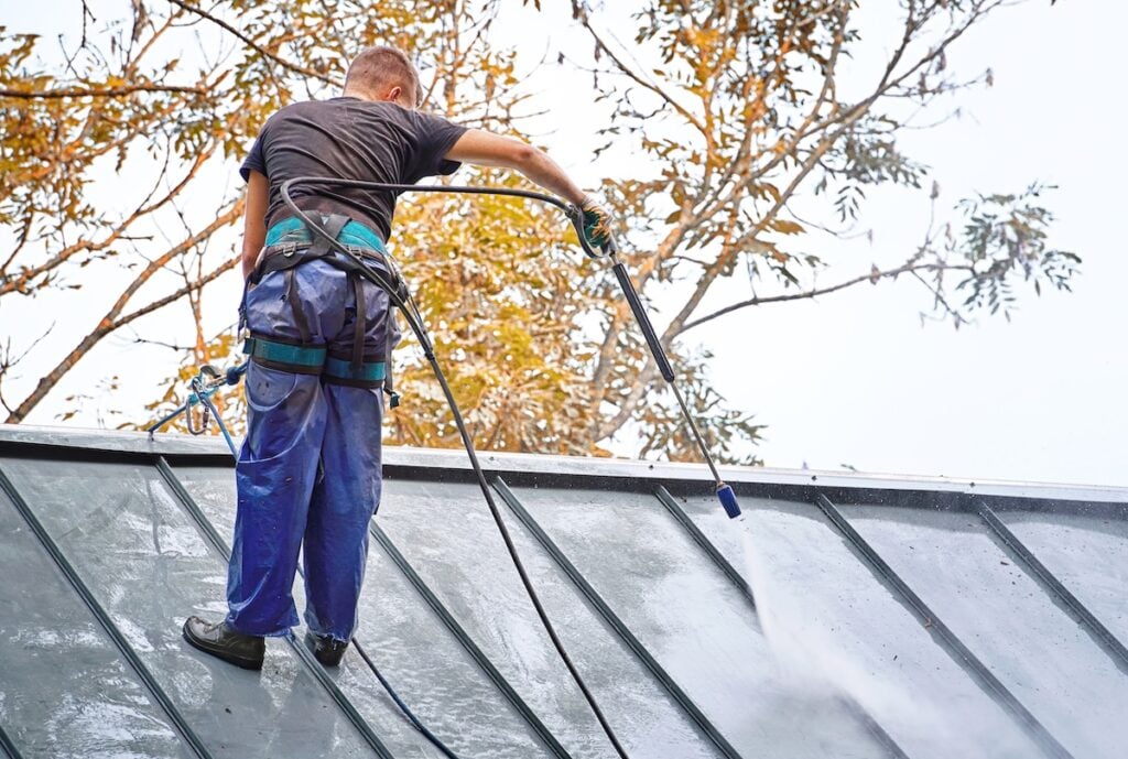 A person wearing safety gear stands on a metal roof, using a pressure washer to clean it. Trees with autumn leaves are visible in the background against a bright sky.