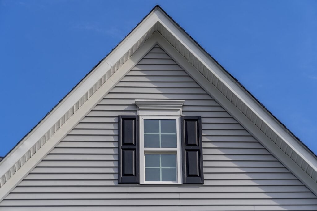 Close-up of the upper part of a house with gray vinyl siding, a triangular gable roof, and a small window with white trim and black shutters against a clear blue sky.