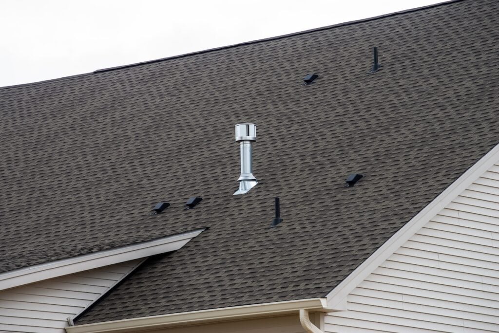 A steeply pitched house roof with brown shingles, a silver metal vent pipe, and multiple black snow guards installed in rows to prevent snow from sliding off. The house has light beige siding.