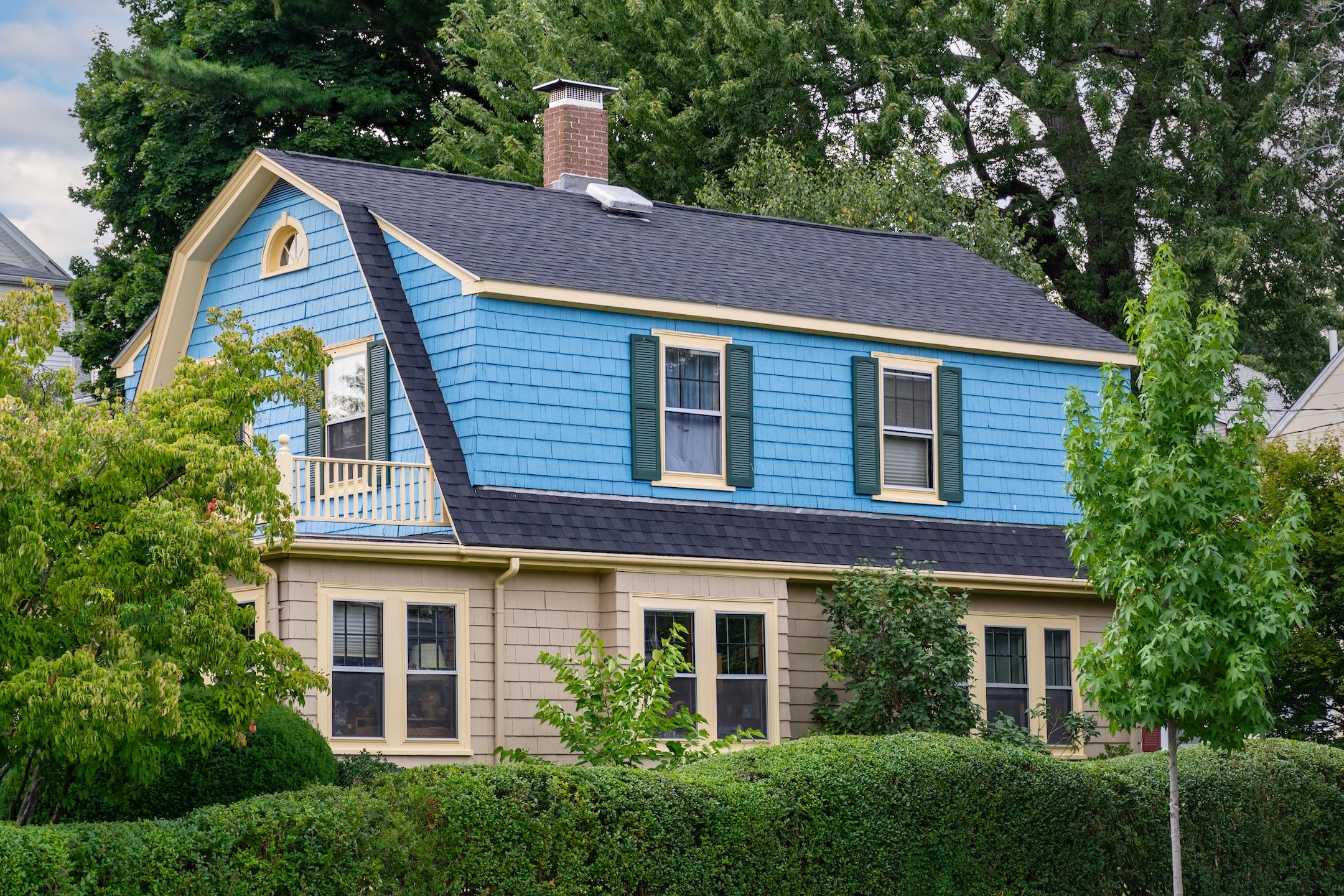 A two-story house with blue siding above and tan below, white trim, green shutters, and surrounded by trees and bushes. The home features a dark gambrel roof and a small balcony.