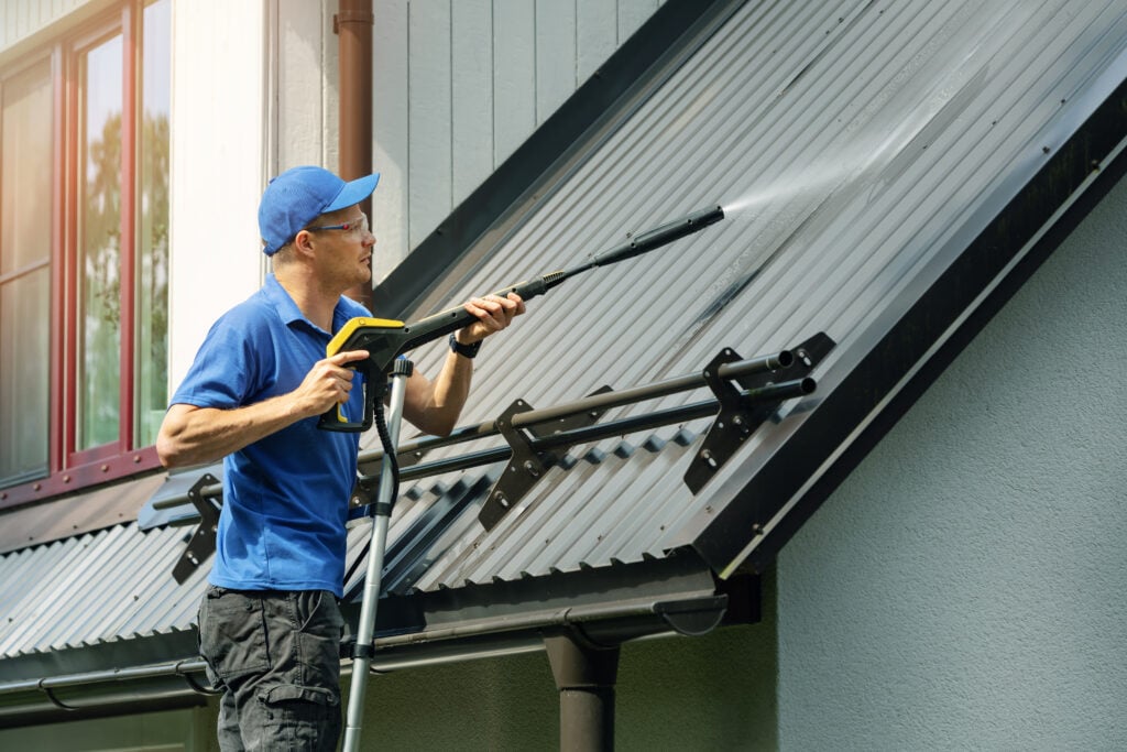 A person in a blue shirt and cap uses a pressure washer to clean a gray metal roof on a house, standing on the ground and aiming the spray upward.