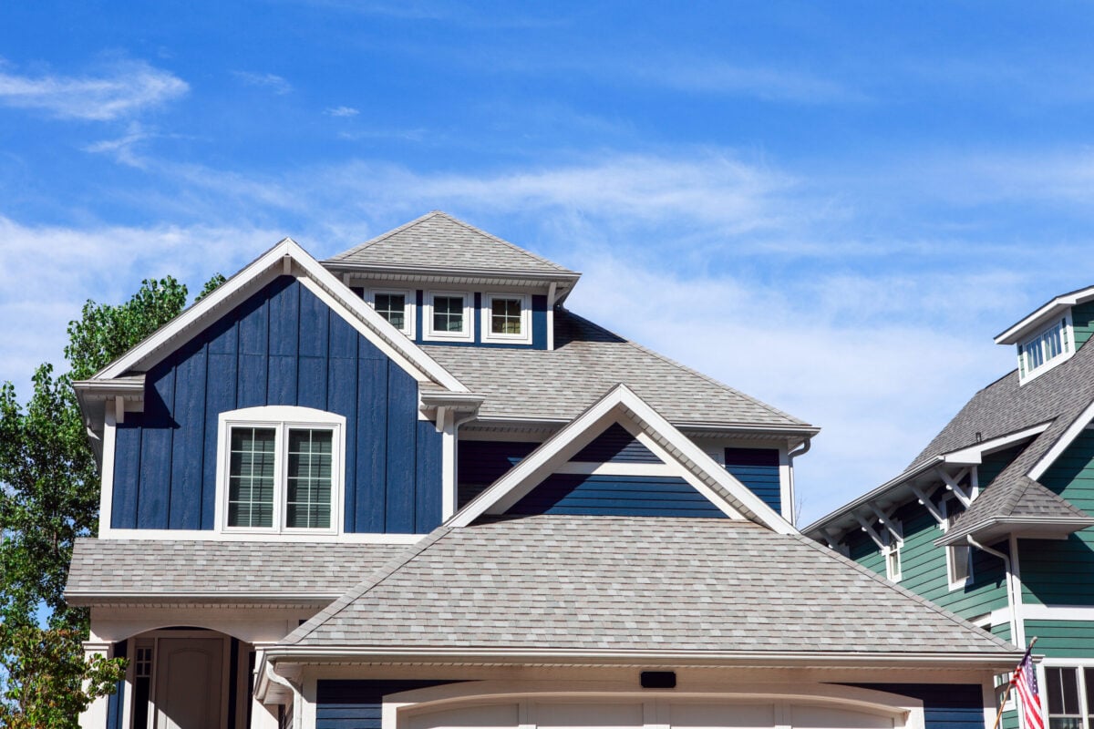 A modern house with dark blue siding and white trim, featuring multiple peaked roofs finished with architectural shingles and several windows, set against a bright blue sky with wispy clouds.