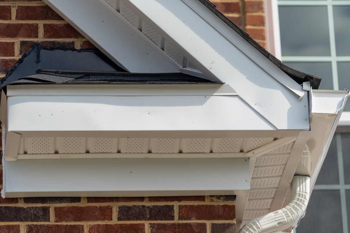 Close-up of a house eave showing white soffit, fascia, gutter, and downspout attached to a brick wall, with a section of black roof shingles and a nearby window.