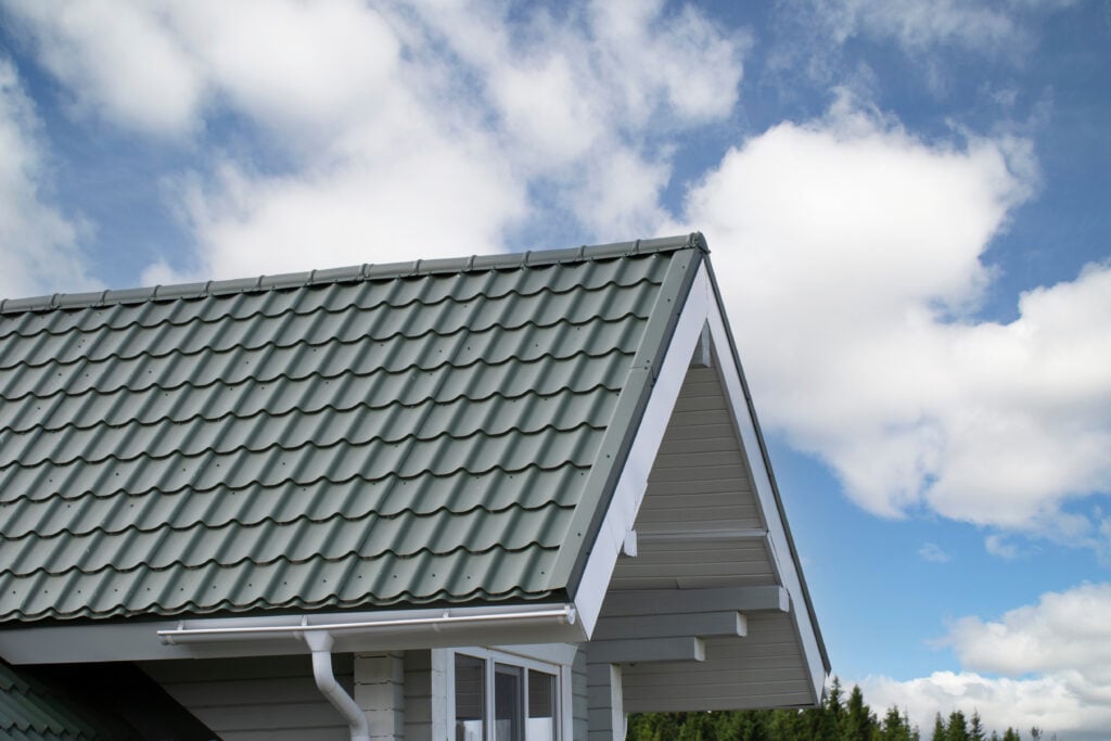 The image shows the roof of a house with green metal roofing tiles, white eaves, and a partly cloudy blue sky in the background.