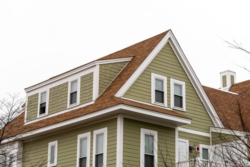 A two-story house with green siding, tan shingle-style accents, white trim, and a brown gabled roof, set against an overcast sky with bare tree branches in the foreground.